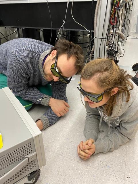A photo showing two young scientists wearing yellow-tinted goggles, lying on the floor of an instrument room staring at a screen in front of them